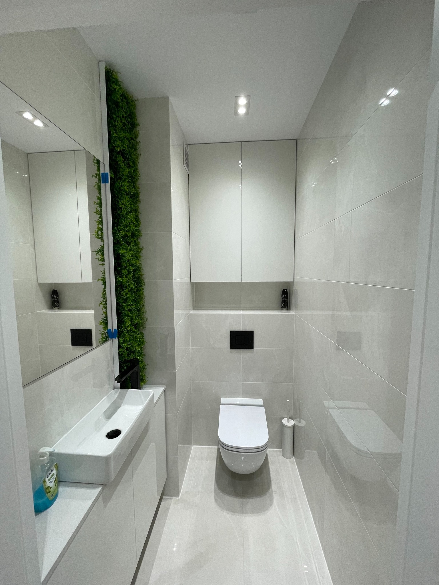 Nowoczesna łazienka z białymi płytkami, wiszącą toaletą i umywalką nablatową, pionowa ściana zieleni obok lustra, minimalistyczny design.