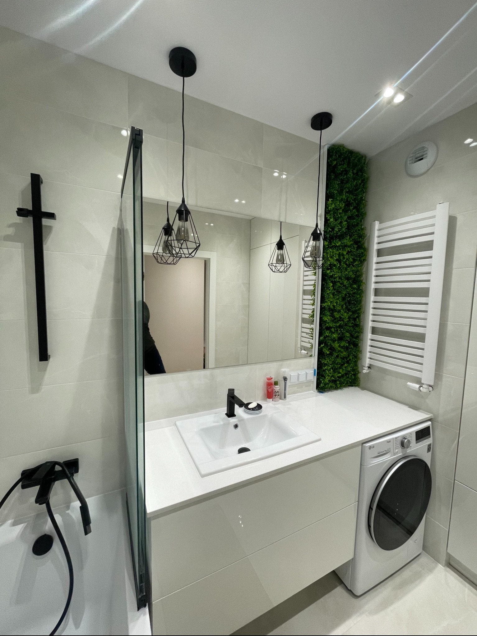 Nowoczesna łazienka z białymi płytkami, umywalką nablatową, czarną armaturą i pionowym panelem z zieleni. Uwagę zwracają geometryczne lampy wiszące i pralka w zabudowie.