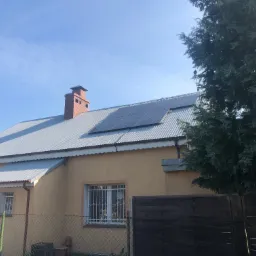 Dom jednorodzinny z zamontowanymi panelami fotowoltaicznymi na metalowym dachu, widoczne okna z kratami i ceglany komin, częściowo zasłonięty drzewem iglastym.