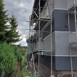 Dwukolorowa elewacja budynku w Malborku, z szarymi pasami oddzielonymi białymi liniami, w trakcie prac wykończeniowych, widoczne rusztowania i pracownik w dolnej części kadru.