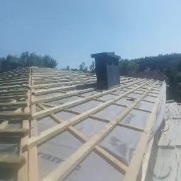 Konstrukcja drewniana dachu z kominem w trakcie prac, widoczna membrana dachowa z nadrukiem, błękitne niebo w tle.