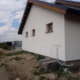 Świeżo otynkowana, biała ściana domu jednorodzinnego z trzema oknami, widoczny fragment dachu z ciemną dachówką, na ziemi gruz i resztki styropianu, w tle blaszany garaż.