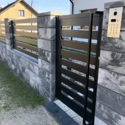 Nowoczesna, pozioma brama wjazdowa z czarnymi, metalowymi szczeblami, osadzona w szarym, betonowym murze z elementami dekoracyjnymi, z widocznym domofonem na słupku.