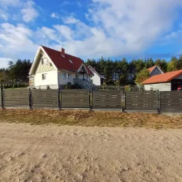 Nowoczesne poziome przęsła w szarym kolorze, tworzące ogrodzenie posesji z domem jednorodzinnym i garażem w tle, widok z piaszczystej drogi.