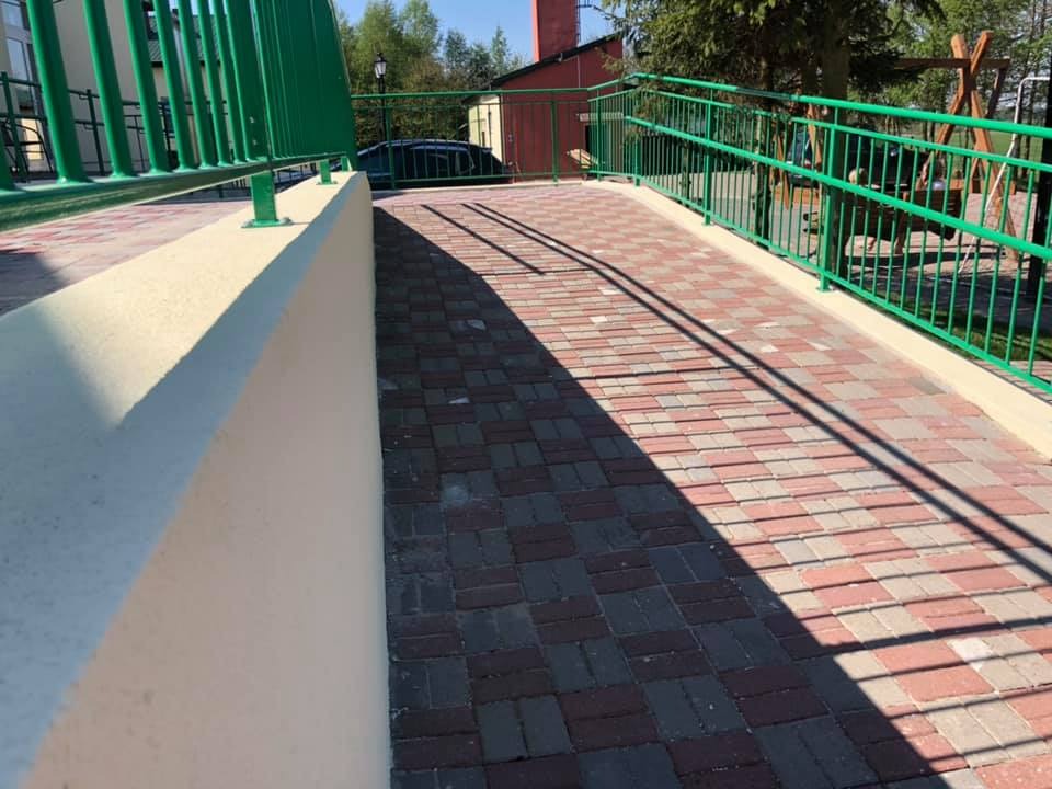 Podjazd z kostki brukowej z zielonymi barierkami, przy budynku z elementami ogrodzenia.