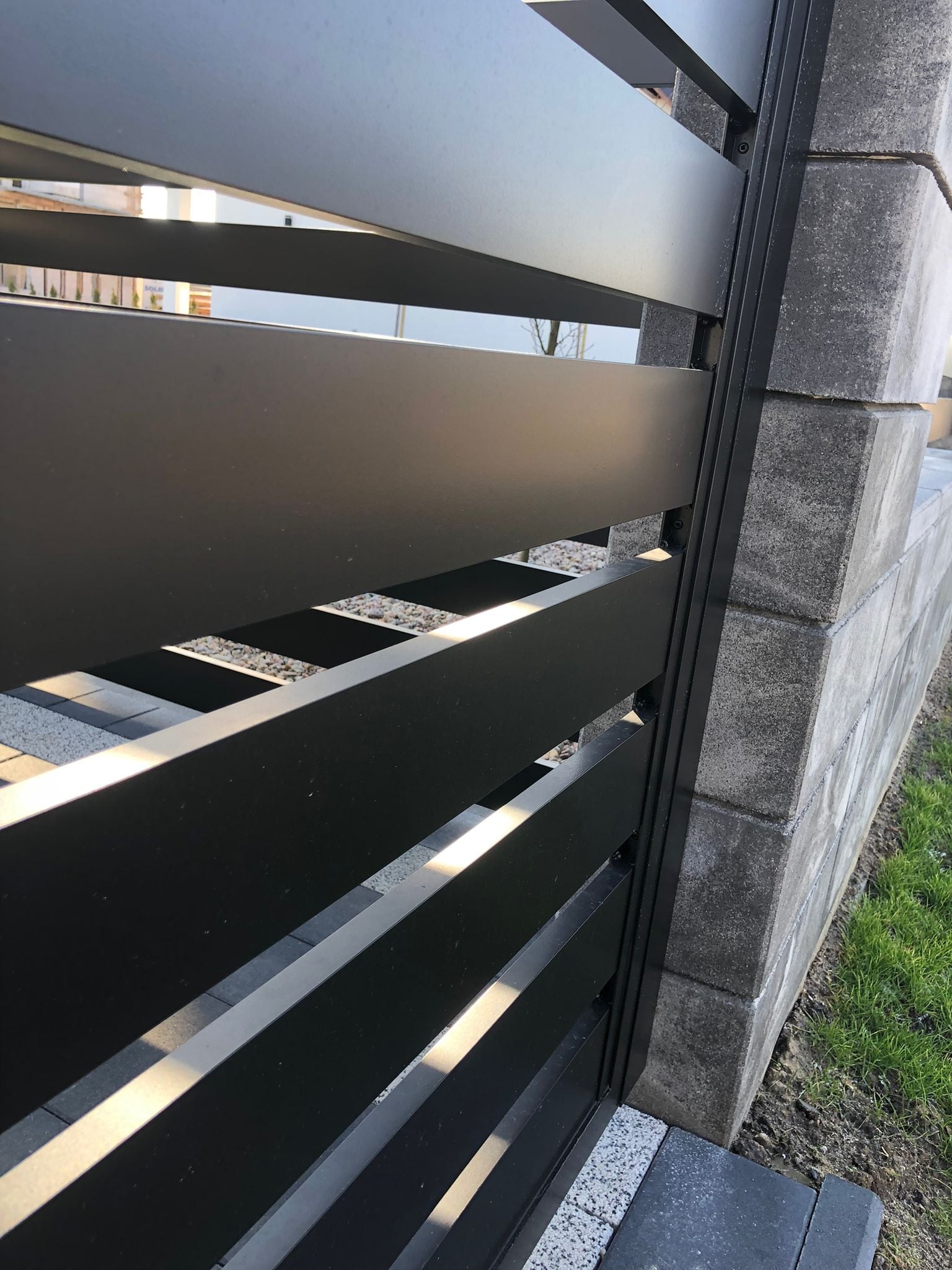 Fragment nowoczesnego, czarnego ogrodzenia z poziomych paneli metalowych, zamocowanego do słupka z szarego betonu architektonicznego, widok z bliska.