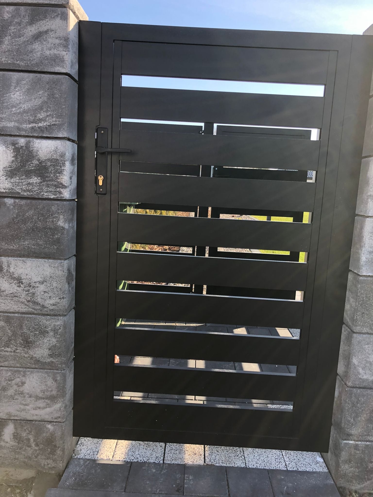 Nowoczesna, czarna brama wjazdowa z poziomymi szczeblami, zamontowana między betonowymi słupkami w kolorze szarości.