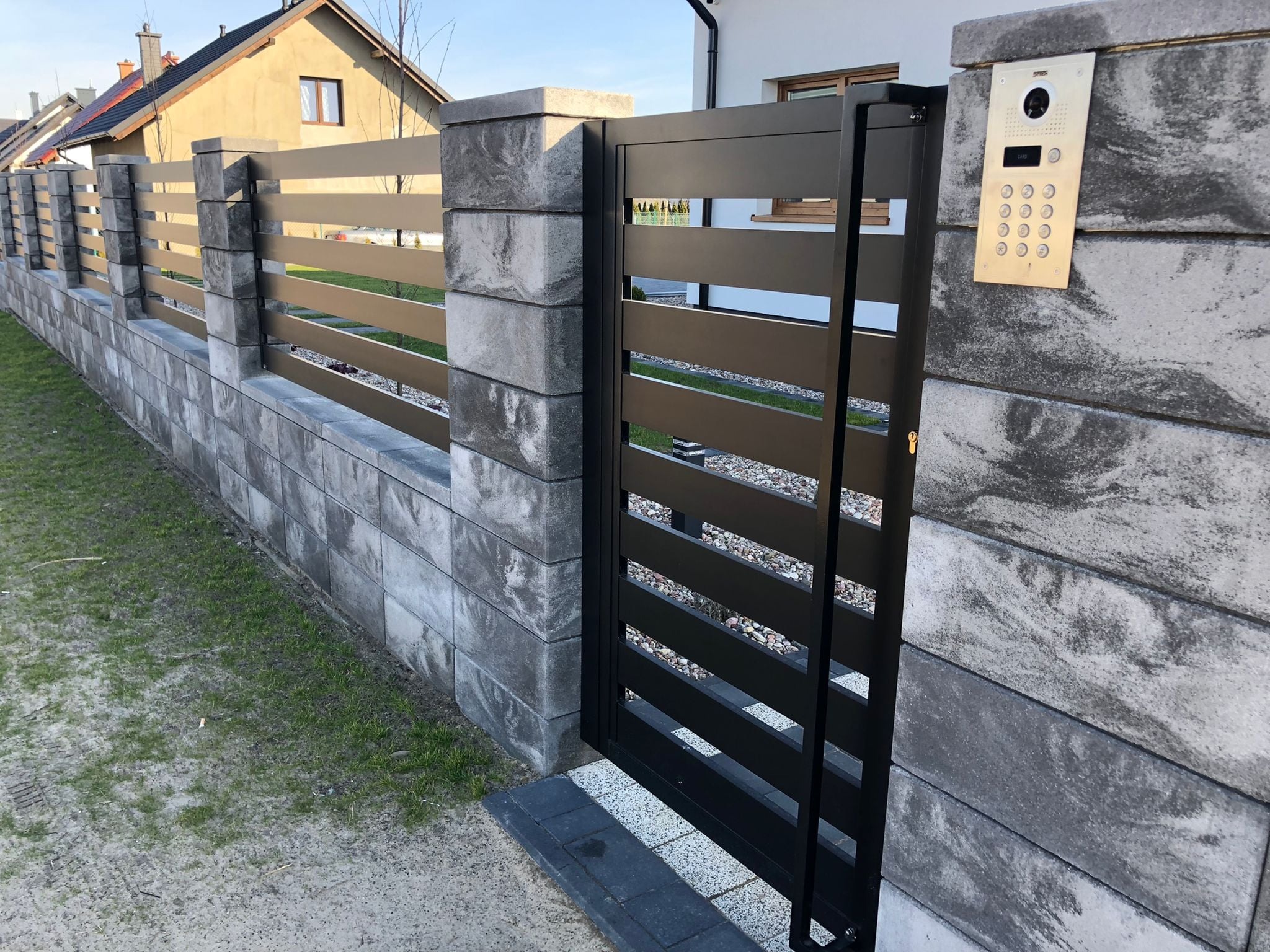 Nowoczesne ogrodzenie z betonowych bloczków i metalowych przęseł oraz czarna furtka z poziomymi szczeblami, z domofonem na słupku.