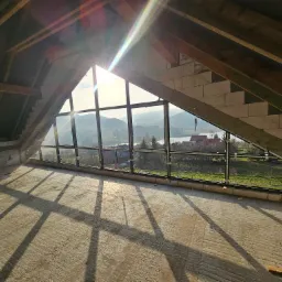 Surowe poddasze z dużym oknem z czarnymi ramami, przez które wpada silne światło słoneczne, widok na zielone wzgórza i jezioro w oddali.