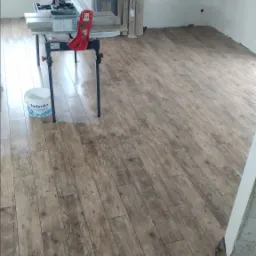 Fragment pokoju z ułożoną podłogą z paneli imitujących drewno; w tle widoczna piła do paneli na stole, paczki paneli i wiadro farby.