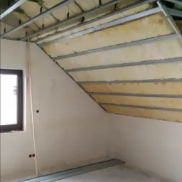 Skośny sufit poddasza w trakcie izolacji wełną mineralną, widoczne metalowe profile konstrukcyjne, fragment okna i ściany z instalacją elektryczną w puszce.