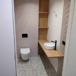 Nowoczesna łazienka z wiszącą białą miską WC, okrągłą umywalką nablatową z czarną armaturą, drewnianym blatem i szafką z półkami, oraz podłogą z lastryko.