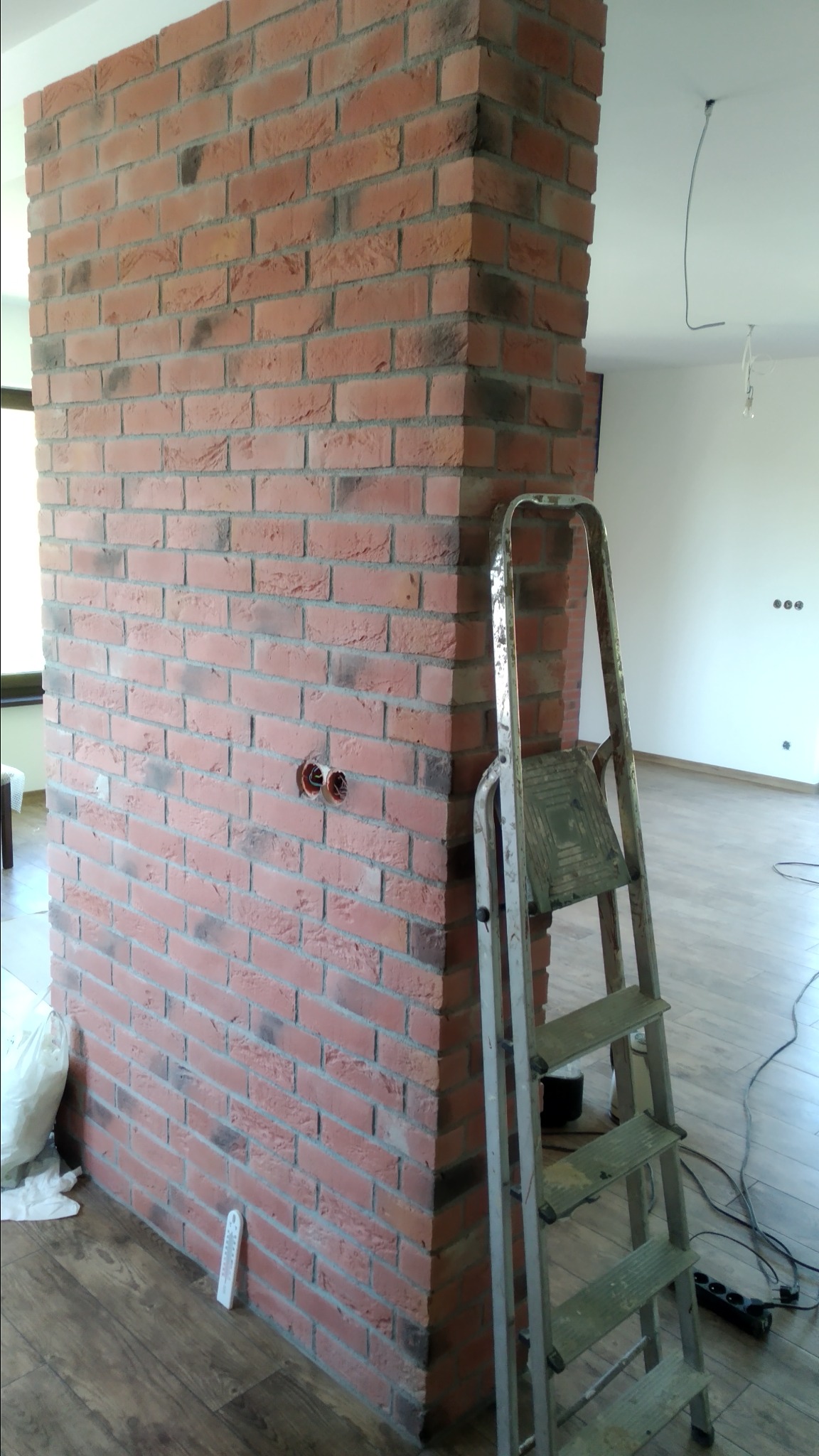 Ceglana kolumna dekoracyjna w surowym wnętrzu, widoczne puszki instalacyjne, drabina aluminiowa i przewody elektryczne na podłodze.