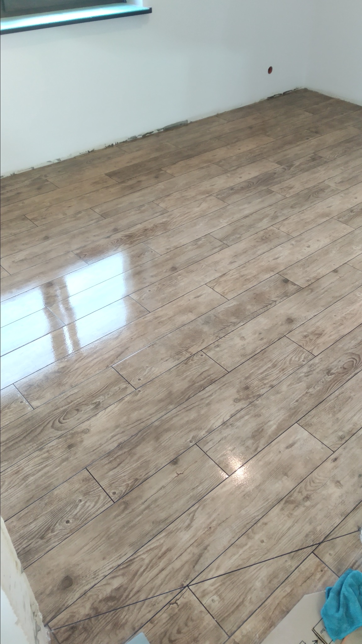 Ułożona podłoga z paneli imitujących drewno, z widocznymi liniami fug, odbijająca światło okna, z fragmentarycznie widocznymi narzędziami i materiałami w rogu pomieszczenia.