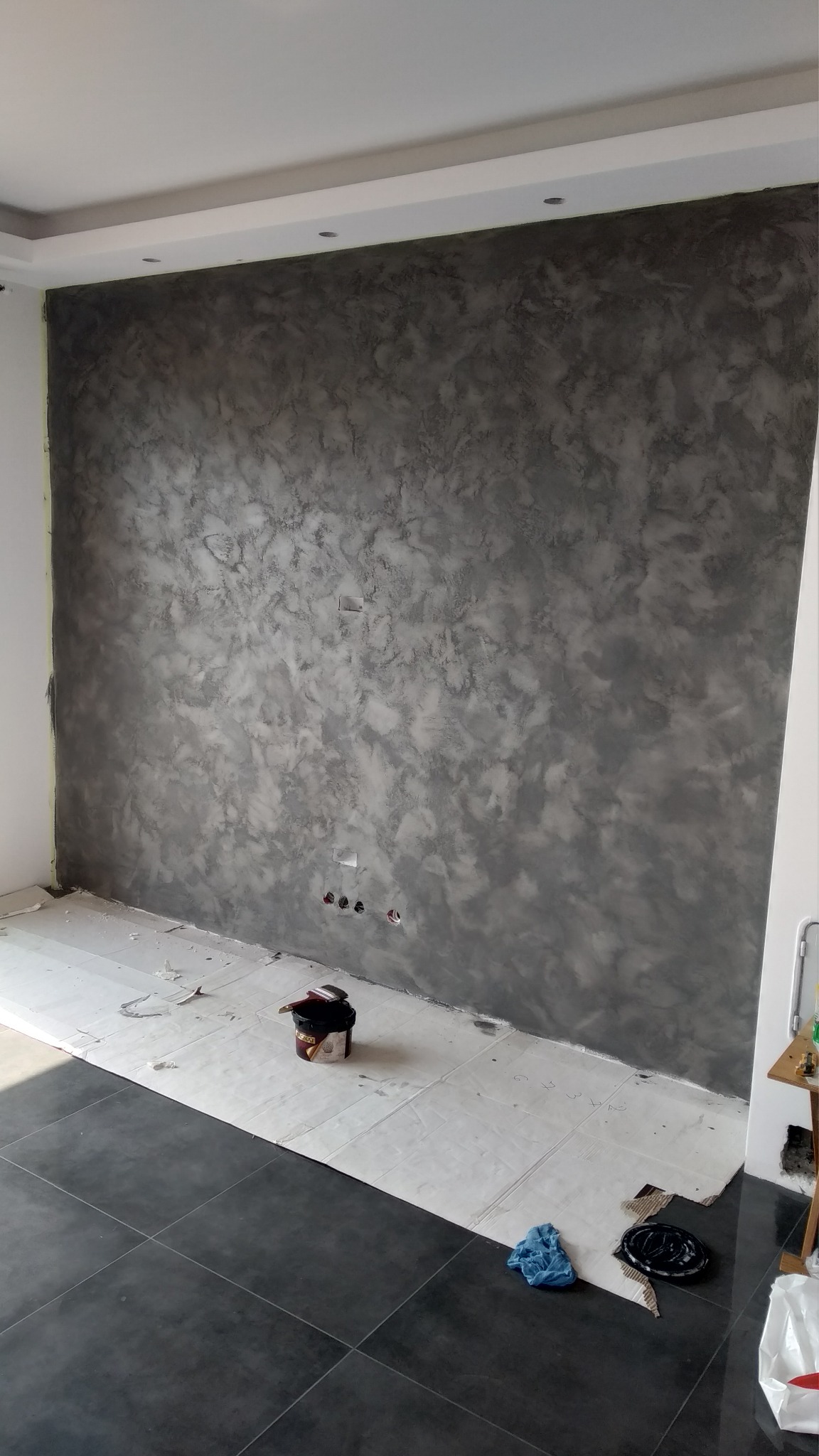 Ściana z efektem dekoracyjnym w odcieniach szarości, zabezpieczona podłoga z widocznym pojemnikiem z farbą i pędzlem, gniazdka elektryczne przed wykończeniem.