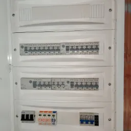 Otwarta, biała skrzynka rozdzielcza z zainstalowanymi bezpiecznikami EATON i ETI, widoczne różne typy wyłączników i zabezpieczeń, ściana z cegły w tle.