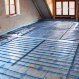 Niebieskie rurki ogrzewania podłogowego, rozłożone równomiernie na srebrnej folii izolacyjnej w surowym wnętrzu poddasza, przygotowane do zalania wylewką.