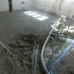 Wnętrze w trakcie wylewania posadzki, widoczne węże doprowadzające materiał i świeżo wylana, jeszcze mokra powierzchnia podłogi, z widocznymi śladami po rozprowadzaniu.