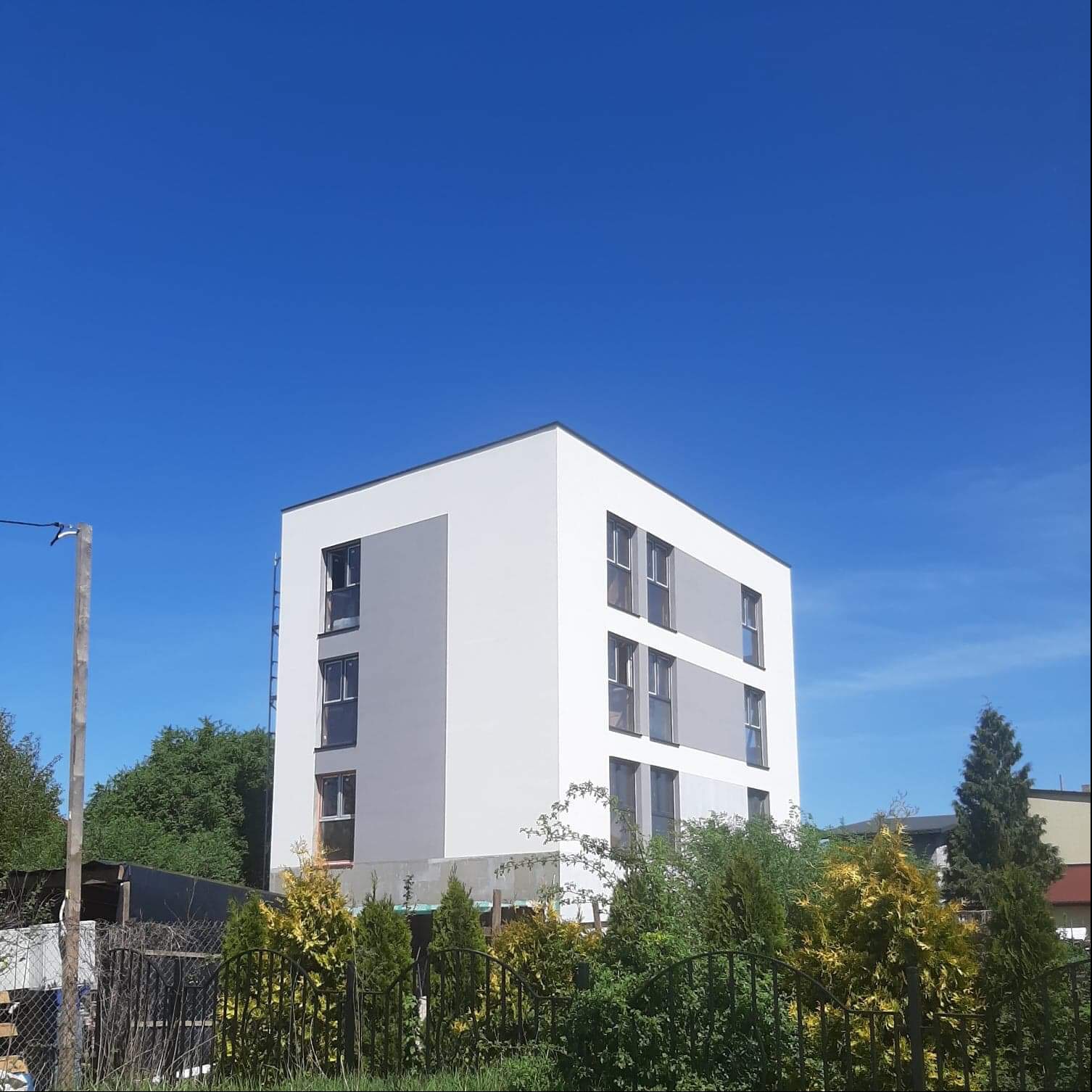 Nowoczesny budynek wielorodzinny z biało-szarą elewacją, widoczny zza ogrodzenia porośniętego zielenią na tle błękitnego nieba.