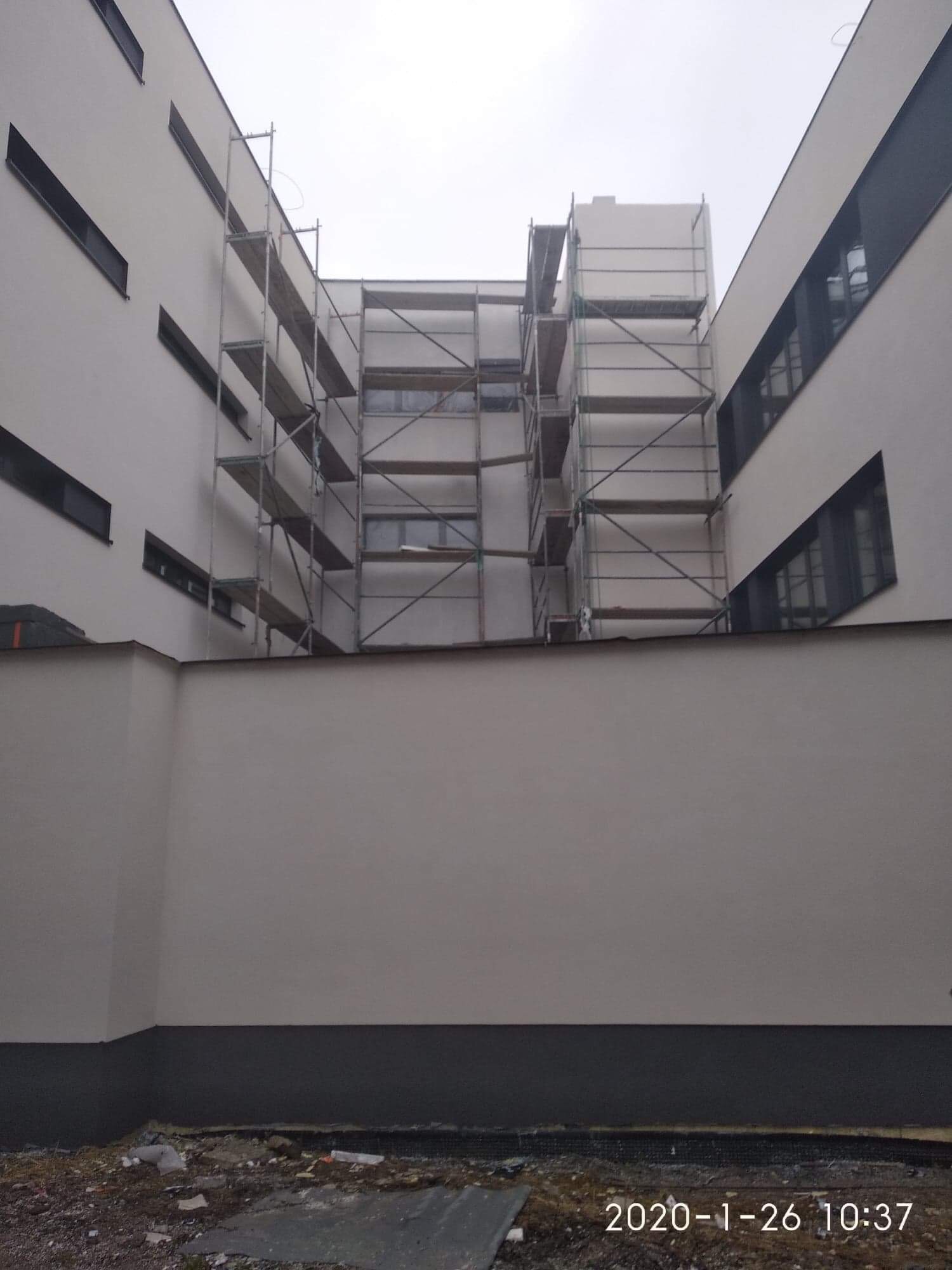 Elewacja budynku w trakcie malowania, widoczne rusztowania na tle białych ścian i rzędów okien, na dole ciemnoszara podmurówka i teren budowy.