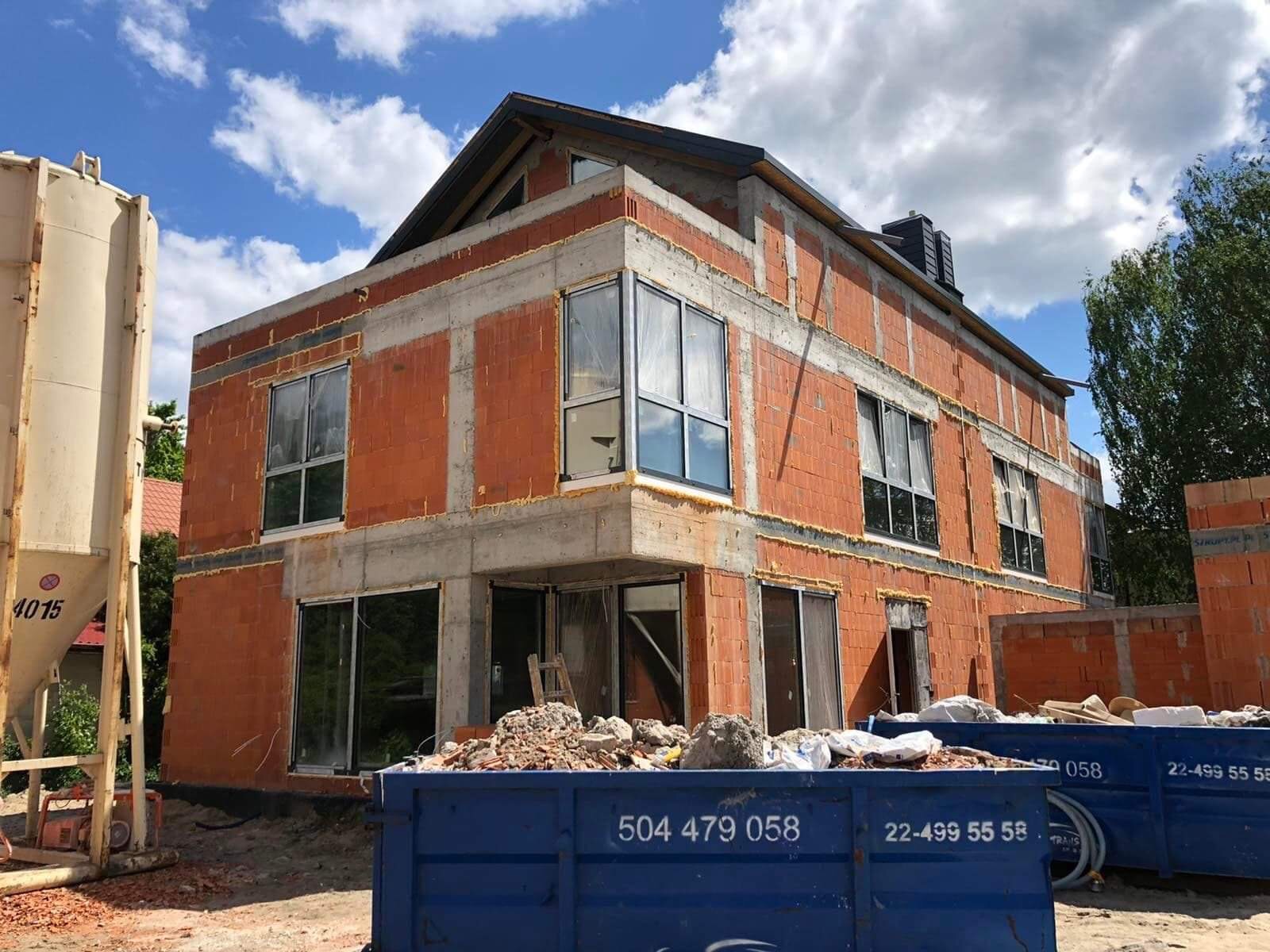 Surowy stan dwupiętrowego domu z czerwonej cegły z betonowymi wzmocnieniami, widoczne okna w metalowych ramach, niebieski kontener na gruz z numerem telefonu na pierwszym planie, silos na zaprawę...