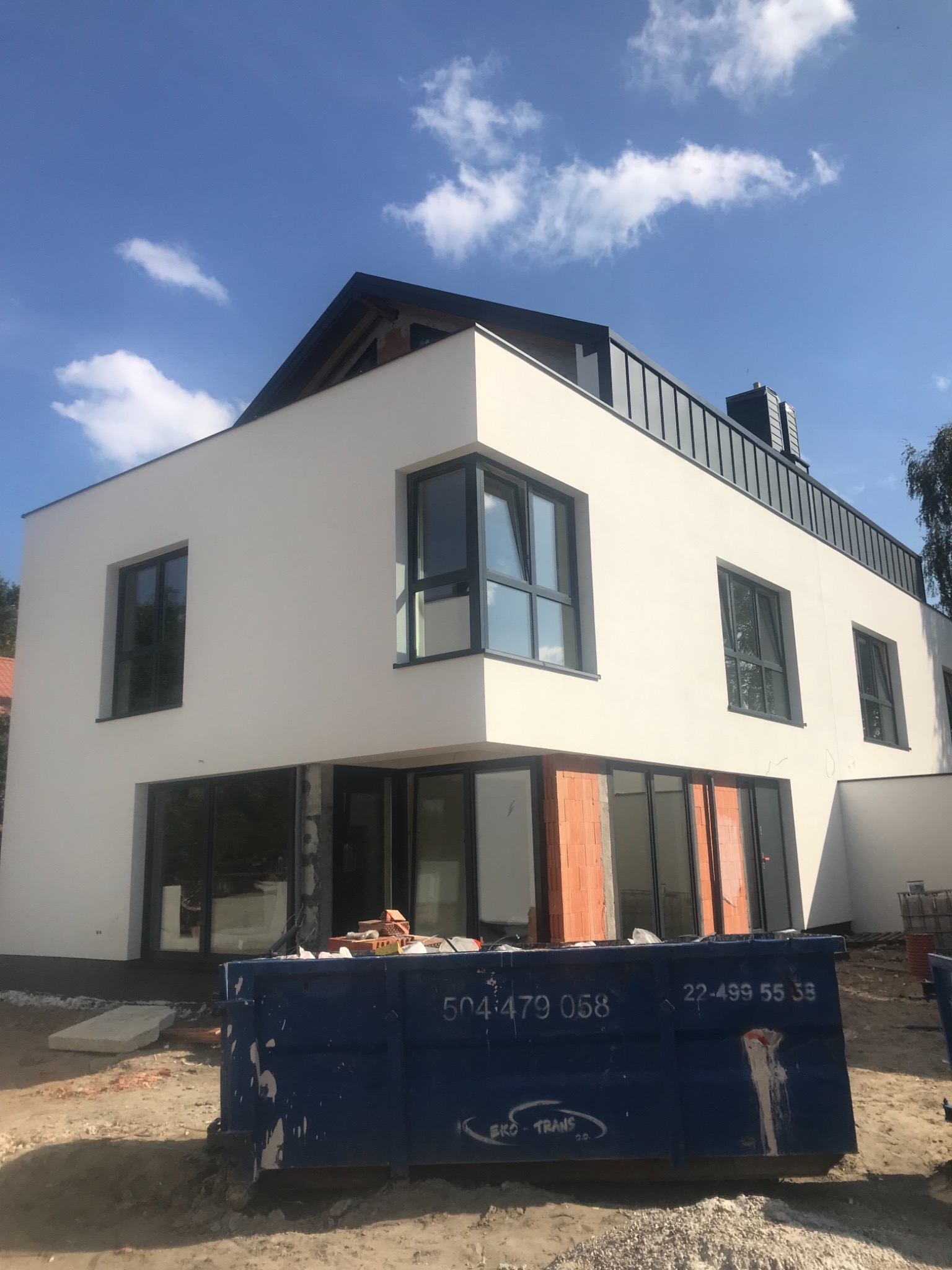 Nowoczesny dom z białą elewacją w trakcie wykończenia, widoczny niebieski kontener na gruz i niezabudowane otwory okienne z ceglanymi ścianami, błękitne niebo z chmurami w tle.