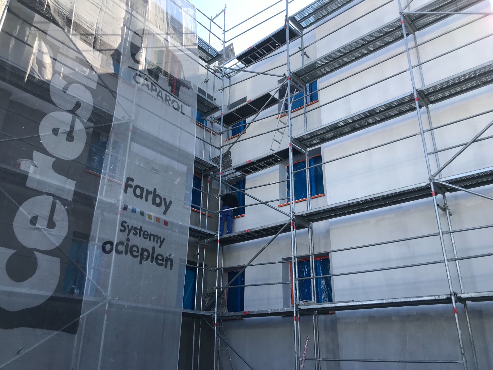 Budynek w trakcie prac elewacyjnych, osłonięty siatką z logo firmy Caparol, widoczne rusztowania i zabezpieczone okna.