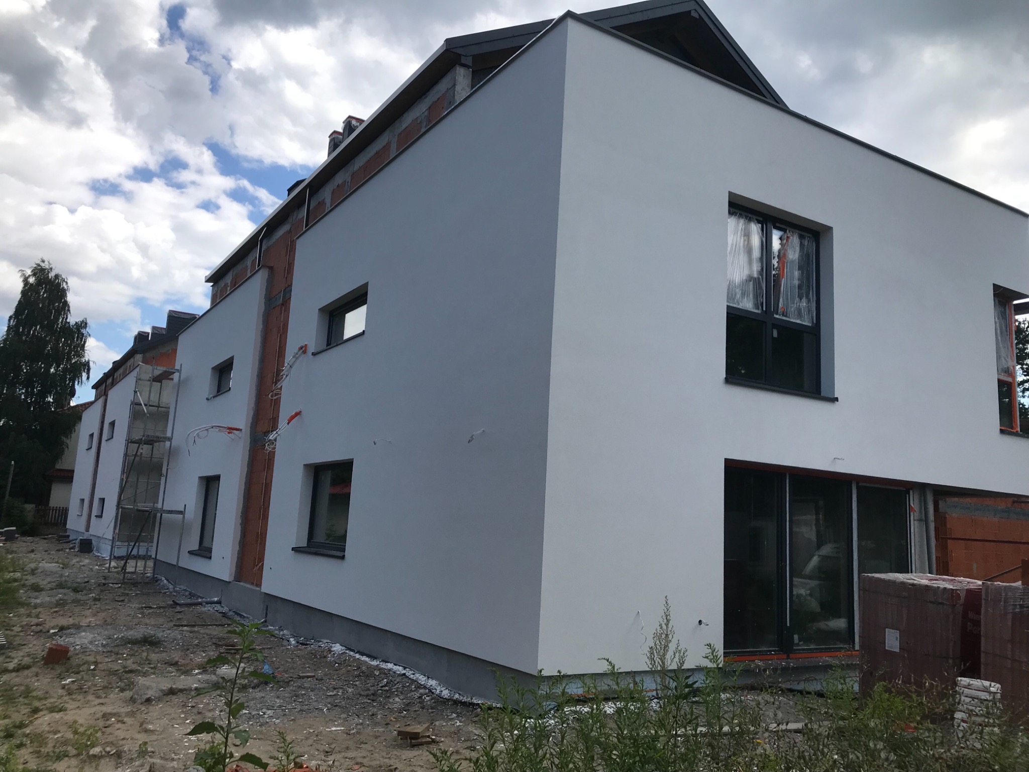 Dwupiętrowy budynek w trakcie prac wykończeniowych elewacji, widoczne rusztowania, częściowo otynkowane ściany i niezabezpieczone okna folią ochronną.