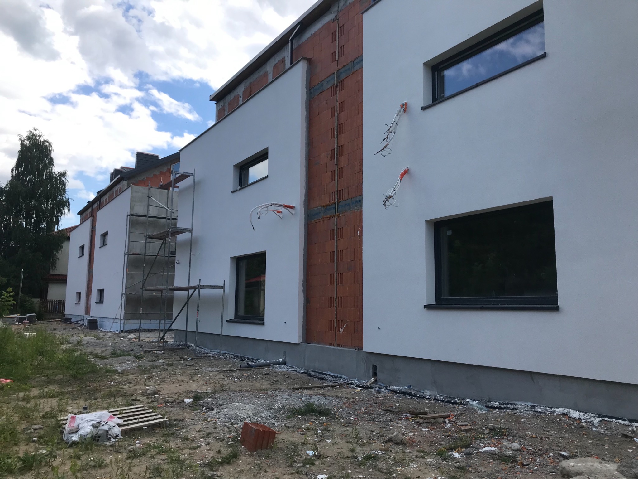 Nowoczesny budynek w trakcie wykańczania elewacji: biała fasada z ceglanymi wstawkami, widoczne kable elektryczne wystające ze ściany, rusztowanie przy bocznej ścianie, niezagospodarowany teren...