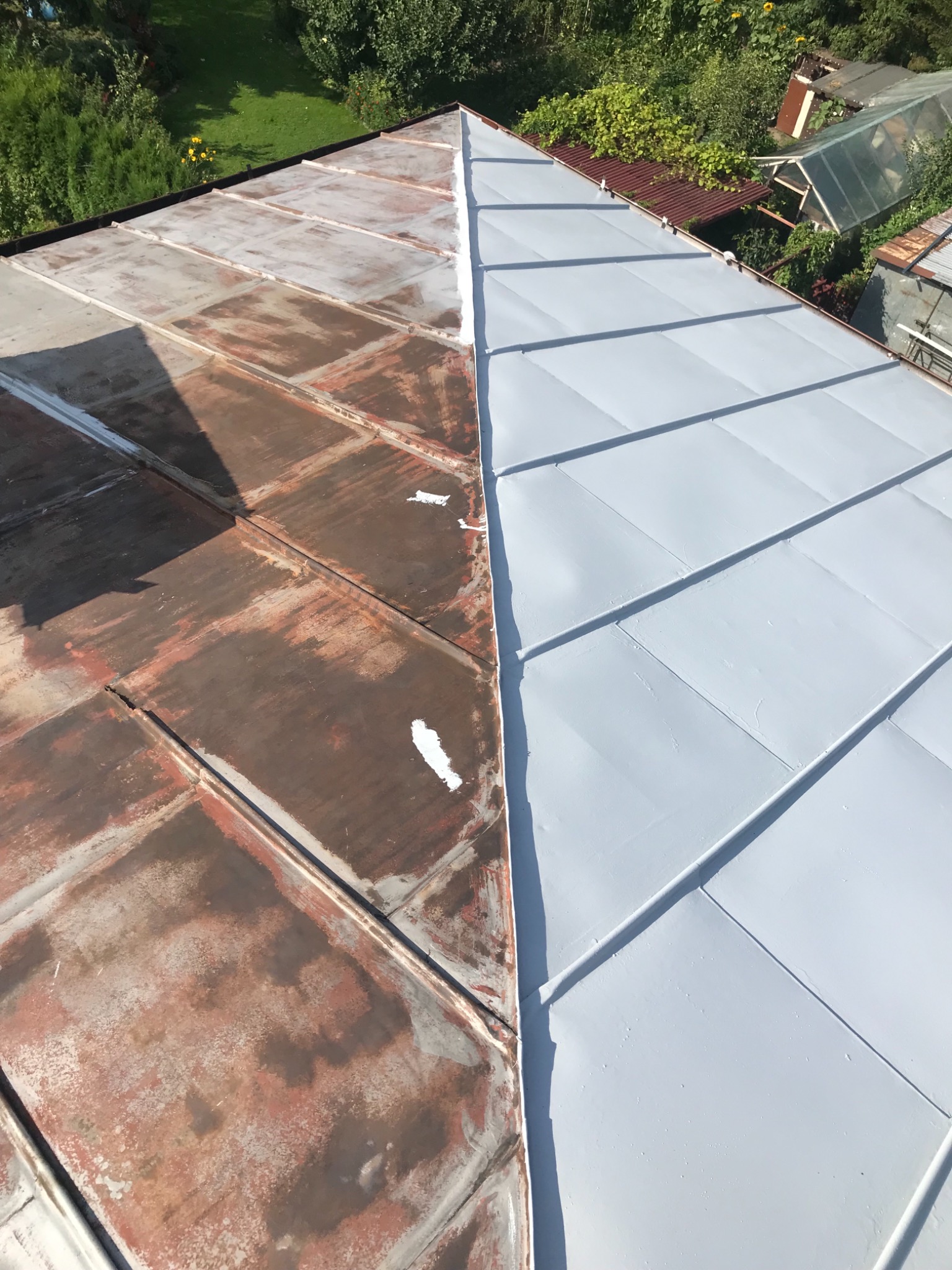 Metalowy dach częściowo odnowiony, z widoczną różnicą między rdzawą, starą powierzchnią a świeżo pomalowaną, jasnoszarą częścią, z zielenią ogrodową w tle.