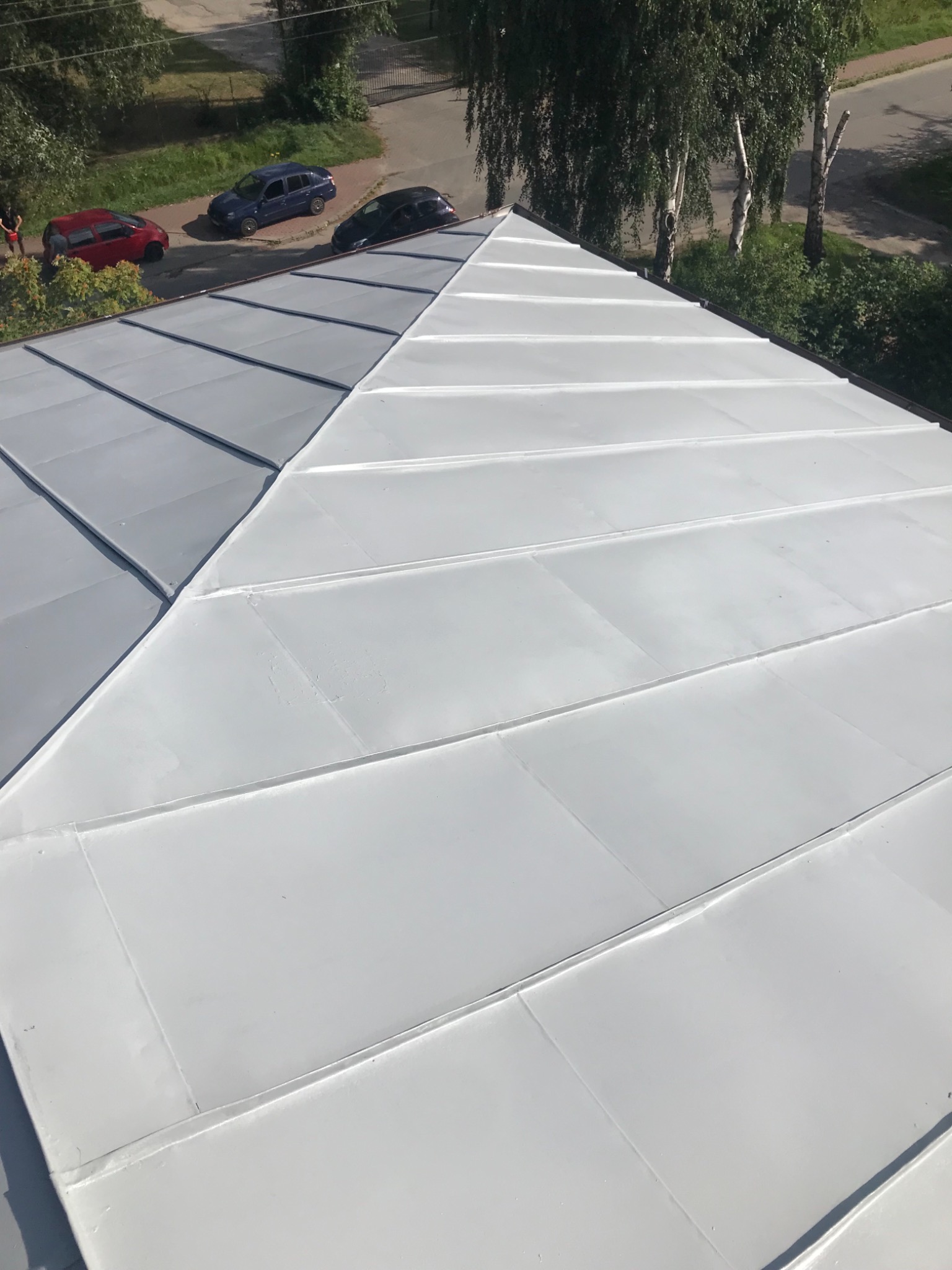 Widok z góry na nowo pomalowany, jasnoszary dach z blachy trapezowej, z widocznymi trzema zaparkowanymi samochodami w tle.