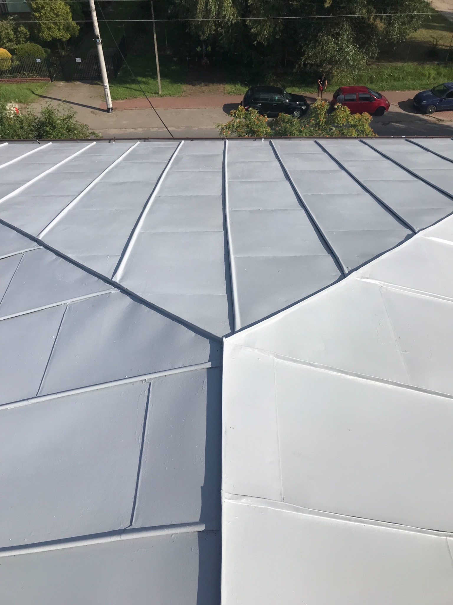 Nowa, metalowa elewacja budynku, częściowo pomalowana na szaro, częściowo na biało, widok z góry, z widocznymi samochodami i zielenią w tle.