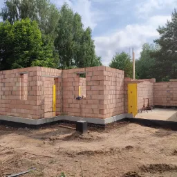 Wznoszenie ścian parteru budynku z czerwonej cegły, widoczne szalunki okienne i drzwiowe, fundament zabezpieczony izolacją, teren budowy z piaskiem i zielenią w tle.