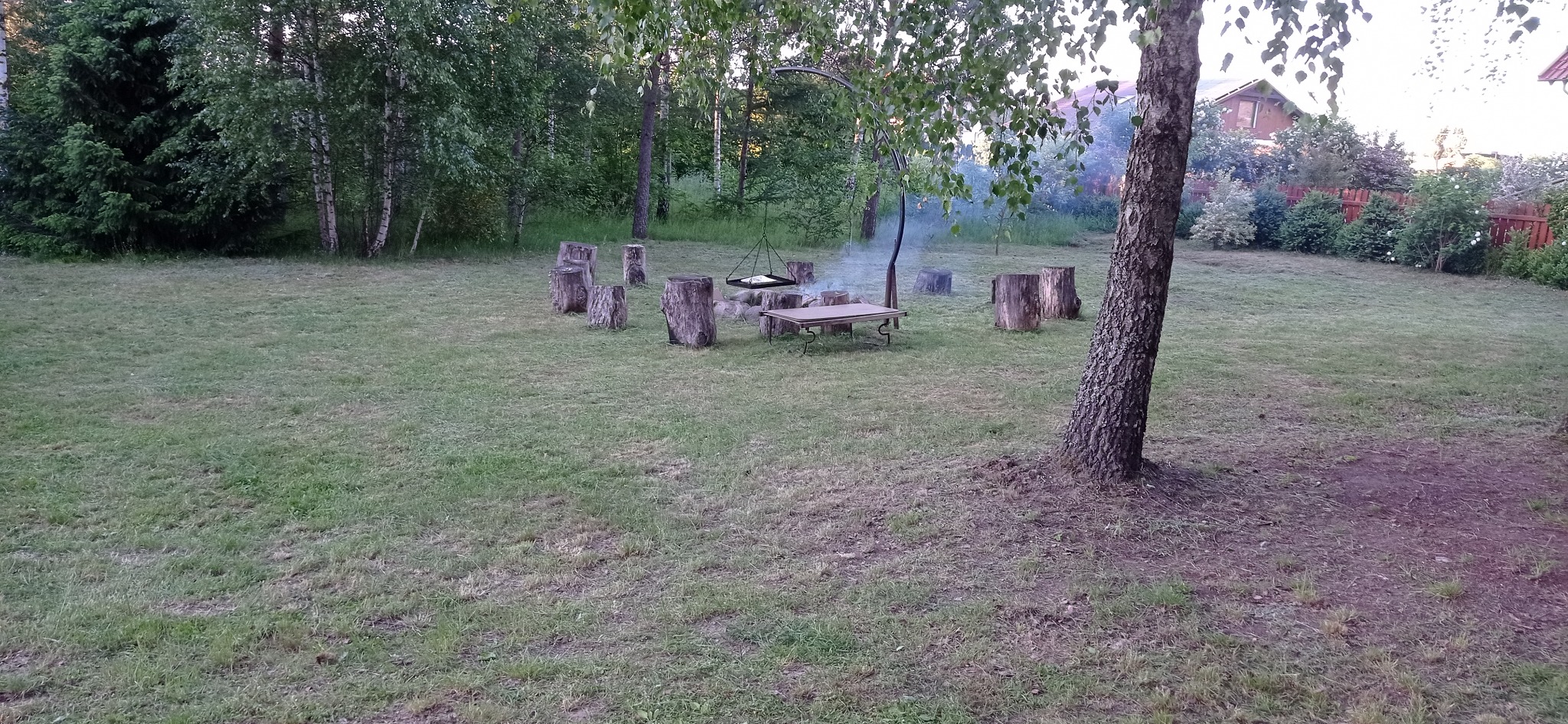 Miejsce na ognisko w ogrodzie z paleniskiem, wiszącym rusztem, stolikiem i siedziskami z pniaków, otoczone trawnikiem i drzewami.