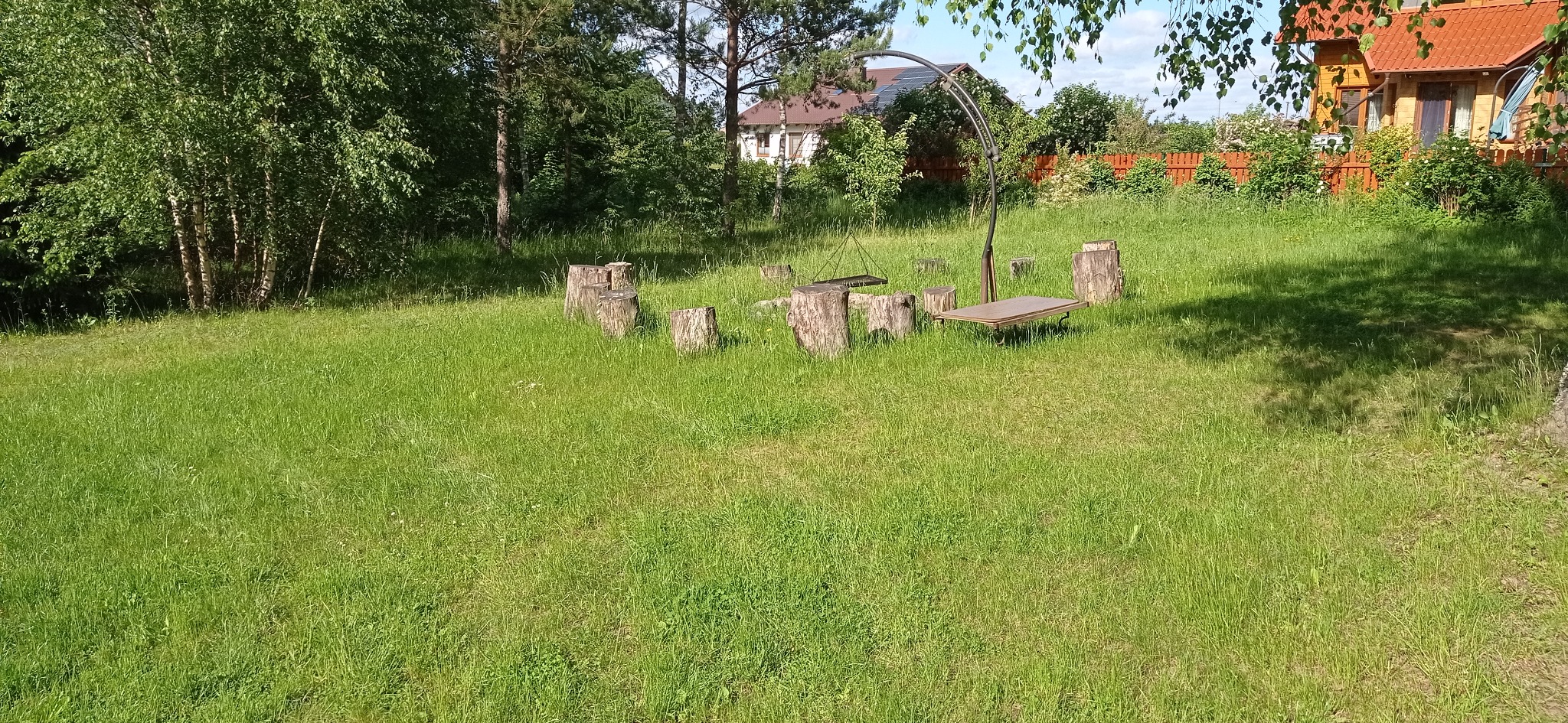 Ogród z miejscem na ognisko: pniaki drzew ustawione w krąg, metalowy grill z rusztem i blatem, wysoka trawa, w tle dom z czerwoną dachówką i drewniany płot.