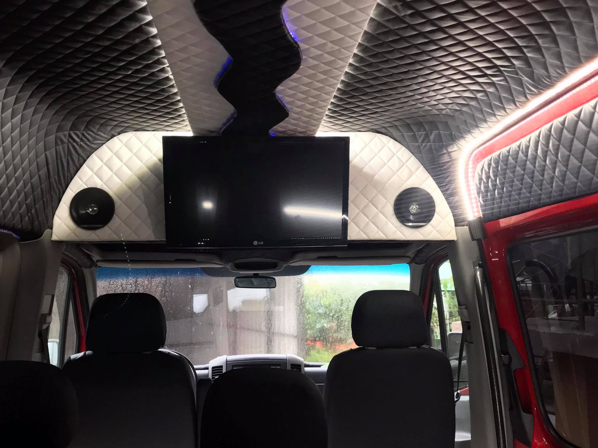 Wnętrze luksusowego busa z pikowaną tapicerką sufitu i ścian, telewizorem LG oraz głośnikami, widok z perspektywy pasażera na przednie siedzenia i szybę z kroplami deszczu.
