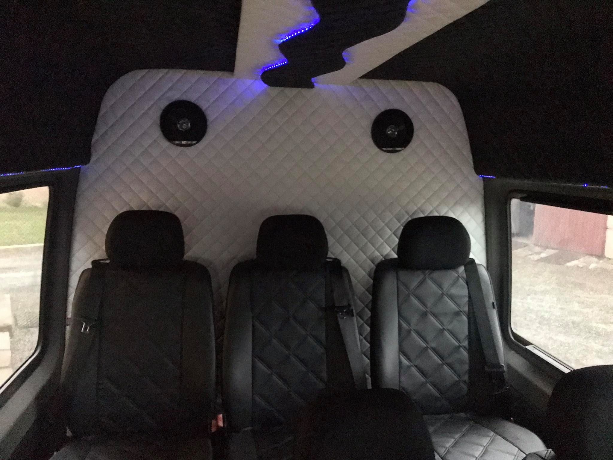 Wnętrze luksusowego busa z trzema skórzanymi fotelami z pikowaniem w romby, białą tapicerką ścian z wbudowanymi głośnikami i niebieskim oświetleniem LED podsufitki.