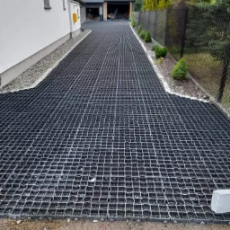 Czarna, ażurowa nawierzchnia drogi dojazdowej z tworzywa sztucznego, obok domu z białym tynkiem i szarym garażem, oddzielona od trawnika z tujami czarną siatką ogrodzeniową.