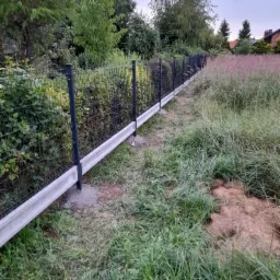 Nowoczesne ogrodzenie panelowe z podmur&oacute;wką betonową, widok z perspektywy trawnika z wysoką trawą, tło z zielenią i domami.