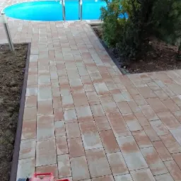 Świeżo ułożona, dwukolorowa kostka brukowa prowadząca do owalnego basenu z drabinką; ubijarka w rogu kadru.