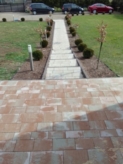 Ułożona kostka brukowa w odcieniach czerwieni i beżu, prowadząca do schodów z jasnych płyt, otoczonych trawnikiem z równo przyciętymi krzewami i małymi drzewkami, w tle zaparkowane samochody.