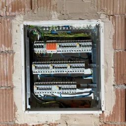 Rozdzielnica elektryczna z zamontowanymi bezpiecznikami i przewodami, umieszczona w surowej ścianie z cegły, widoczne ślady po tynku.