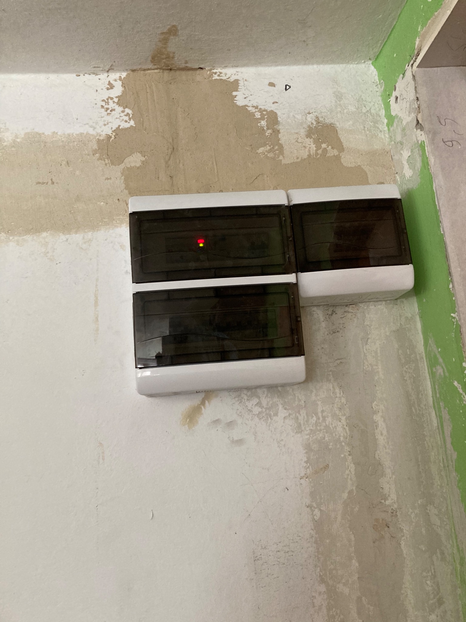 Dwa moduły rozdzielcze natynkowe z białymi obudowami i czarnymi drzwiczkami, zamontowane na nierównej, częściowo otynkowanej ścianie z widocznymi śladami po malowaniu, z czerwoną diodą...