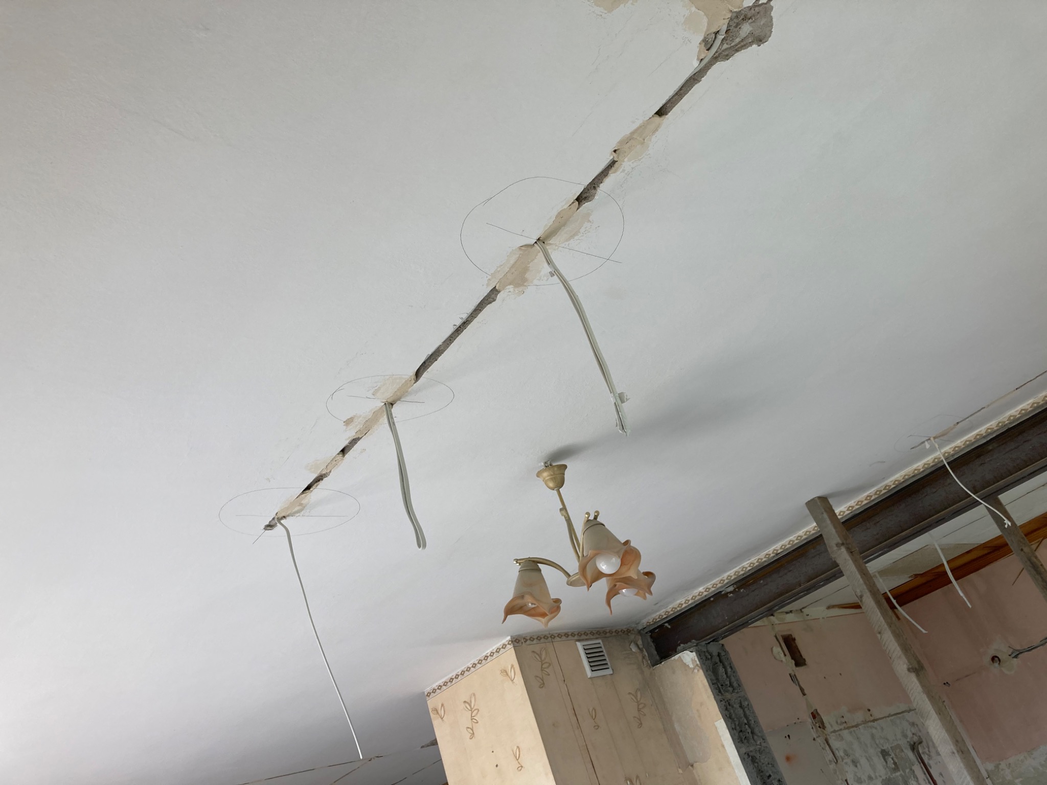 Przygotowanie do montażu oświetlenia sufitowego: wycięte otwory w suficie, wystające przewody elektryczne, widoczna stara instalacja elektryczna i fragment wiszącej lampy.