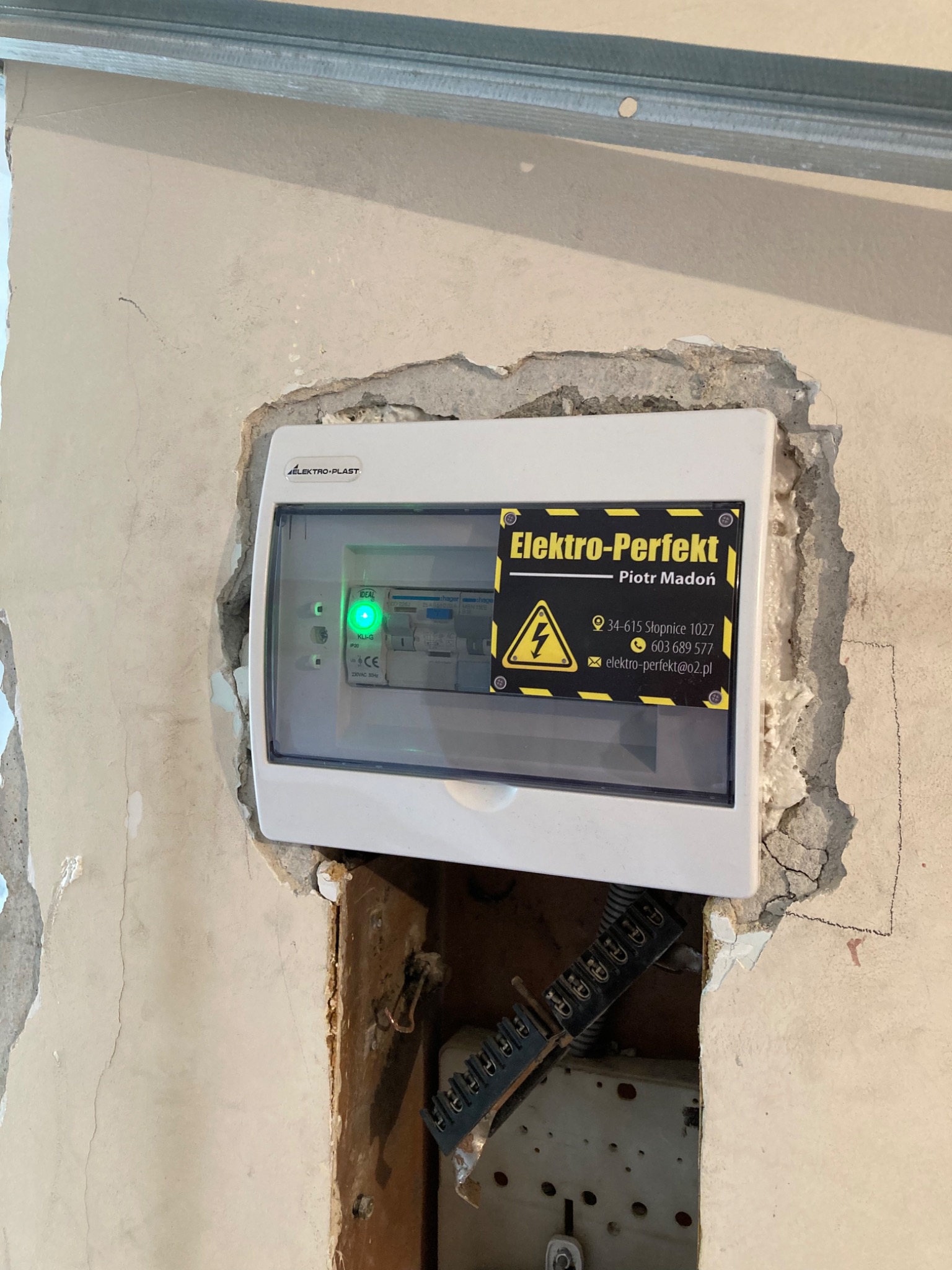 Zamontowana w surowej ścianie skrzynka elektryczna Elektro-Plast z naklejką firmy Elektro-Perfekt, widoczne przewody i elementy instalacji w otworze.