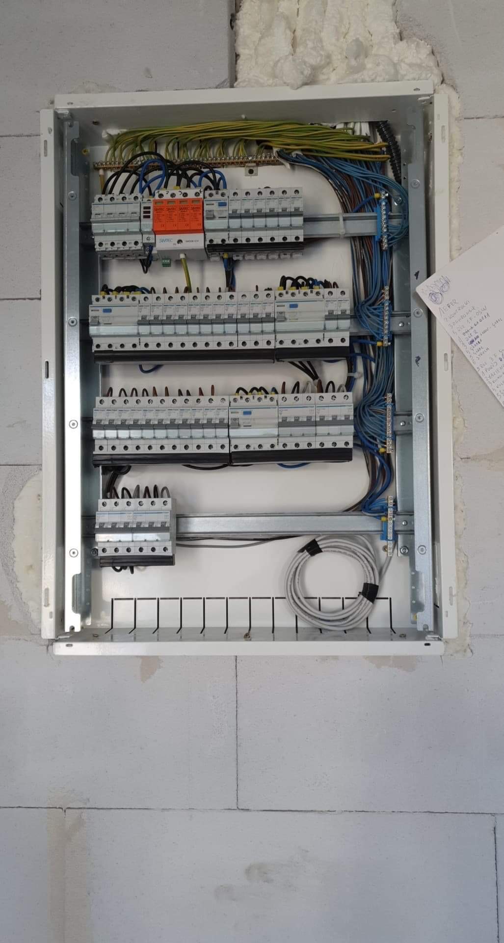 Rozdzielnica elektryczna z zamontowanymi bezpiecznikami i przewodami, widoczna na ścianie z betonu komórkowego, z notatkami przyklejonymi z prawej strony.