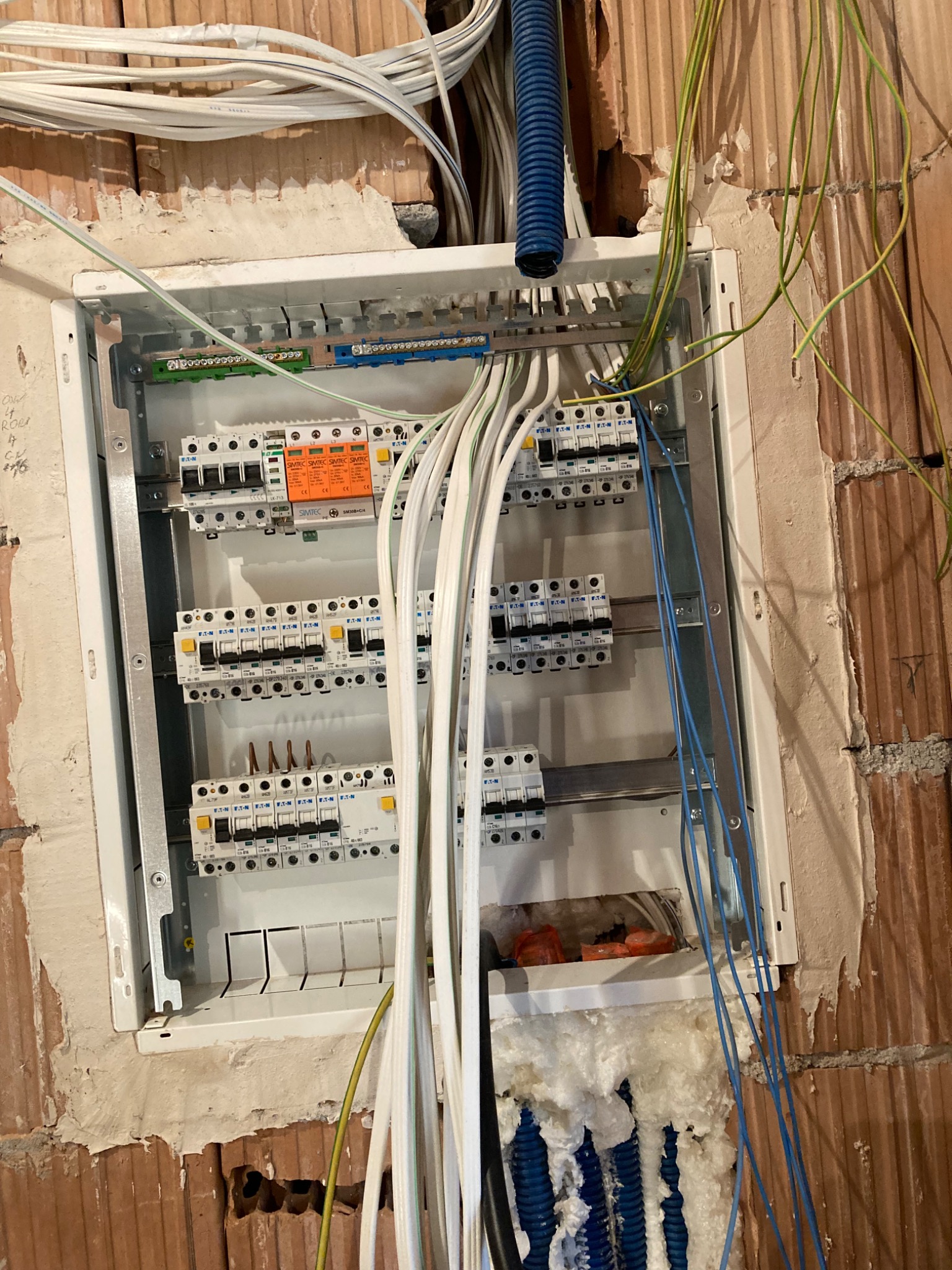 Rozdzielnica elektryczna w trakcie montażu w surowej ścianie z cegły, widoczne przewody w peszlach i bez, zabezpieczenia marki Eaton oraz Simtec, częściowo wypełniona pianką montażową