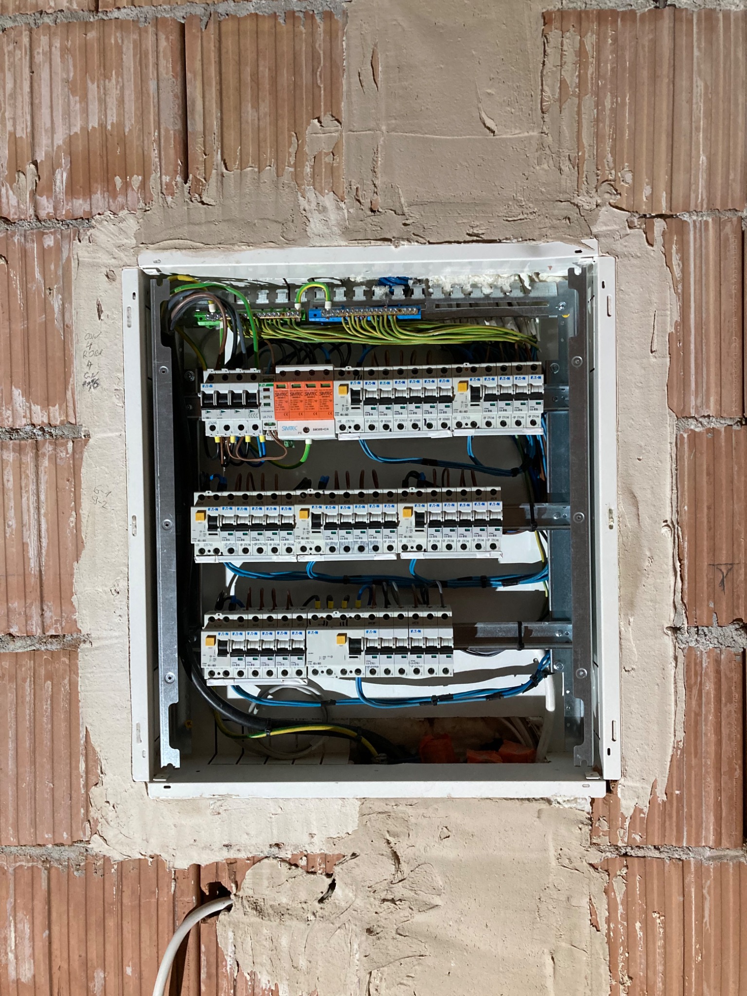 Rozdzielnica elektryczna z zamontowanymi bezpiecznikami i przewodami, umieszczona w surowej ścianie z cegły, widoczne ślady po tynku.
