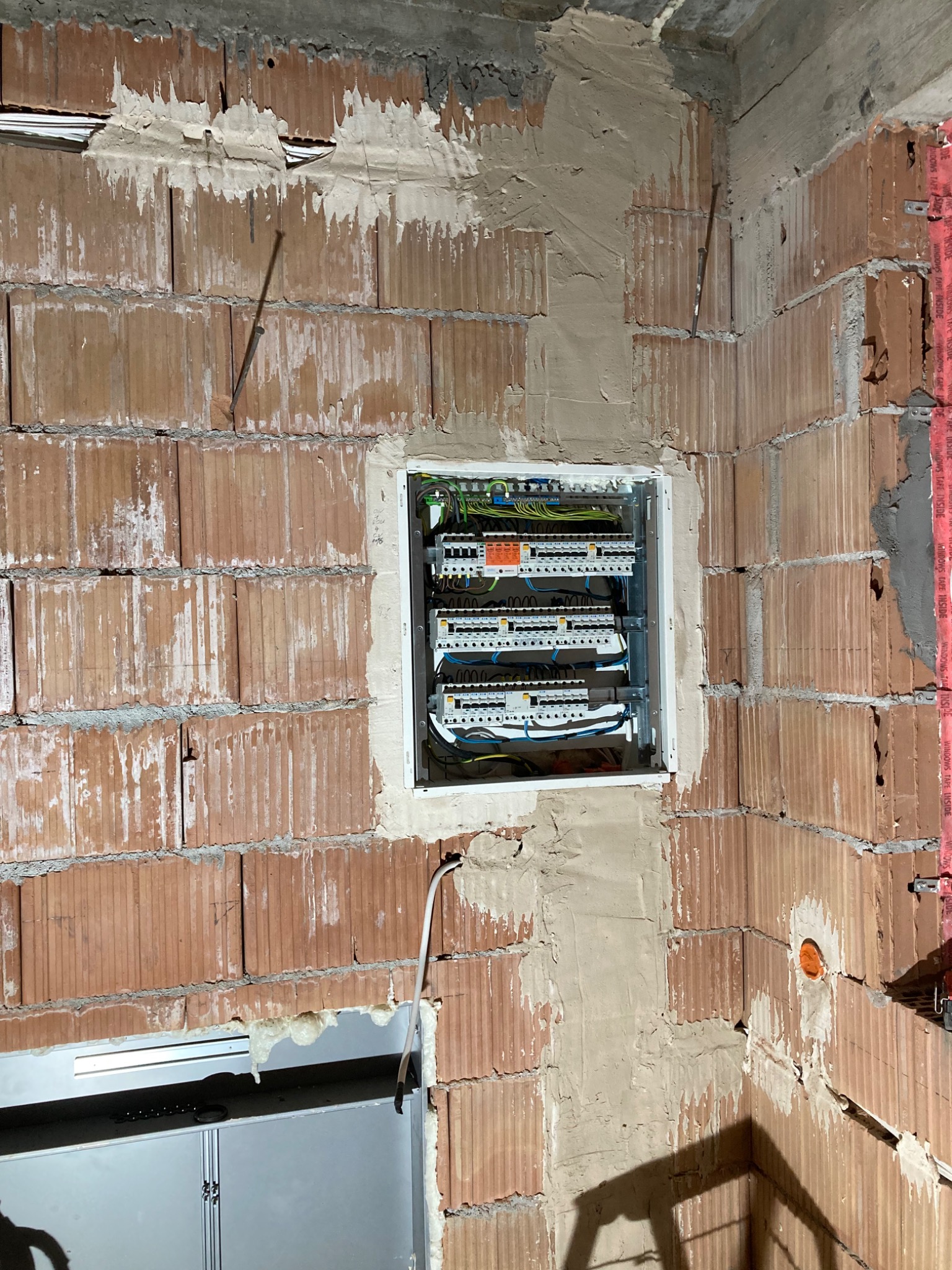 Wnęka w surowej ścianie z cegły dziurawki, w której zamontowano rozdzielnicę elektryczną z widocznymi przewodami i zabezpieczeniami, otoczoną świeżym tynkiem.