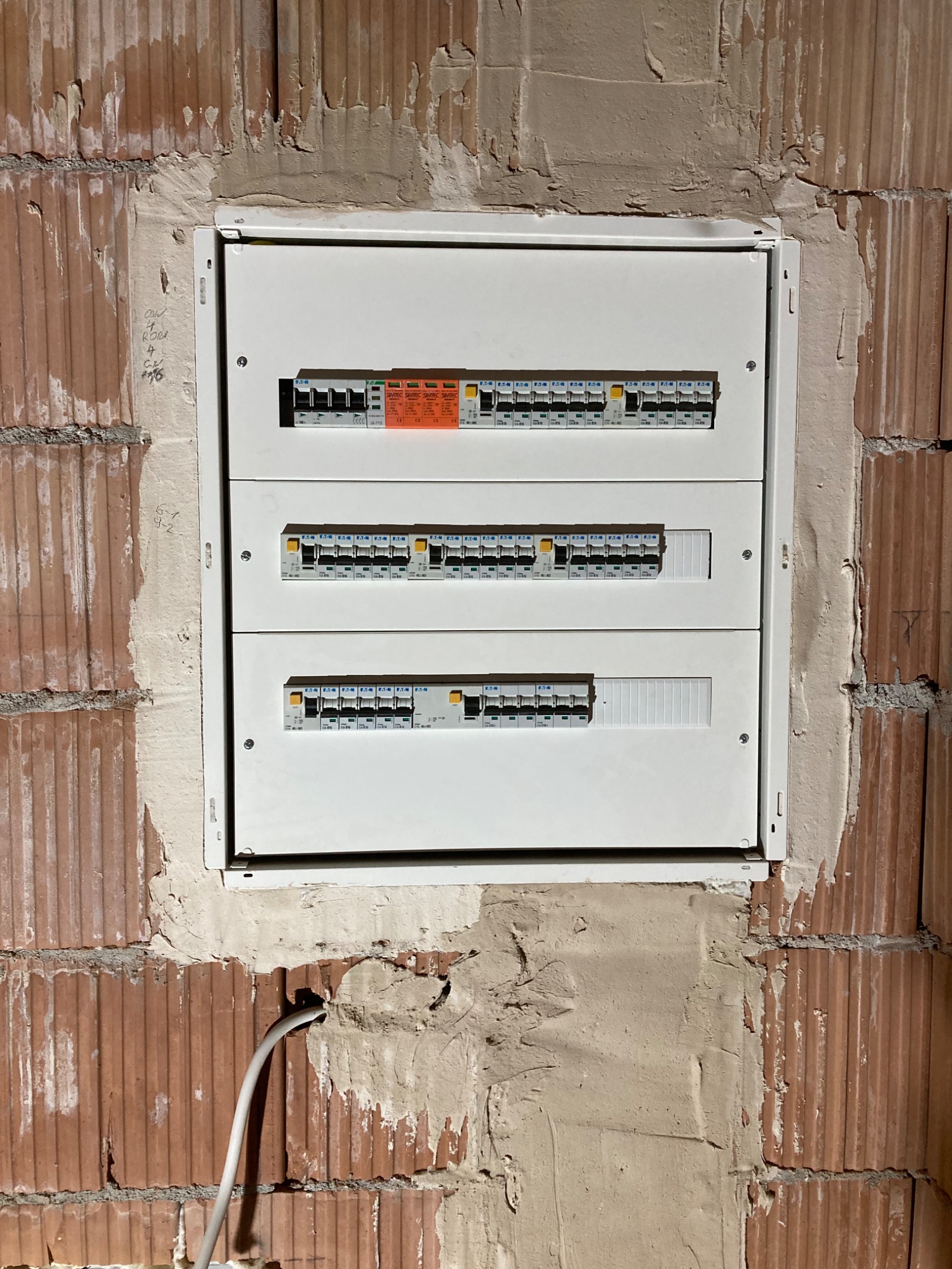 Biała skrzynka rozdzielcza z bezpiecznikami zamontowana w surowej, ceglanej ścianie z widocznymi śladami zaprawy.